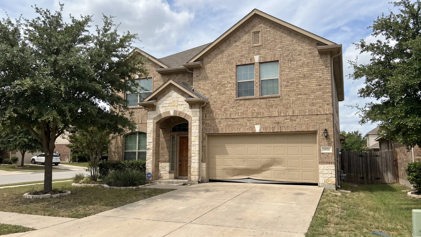 Tan steel garage door with its bottom section buckled inward where a bumper hit it, on a two-story brick home with a stone-trimmed entry in a Central Texas subdivision