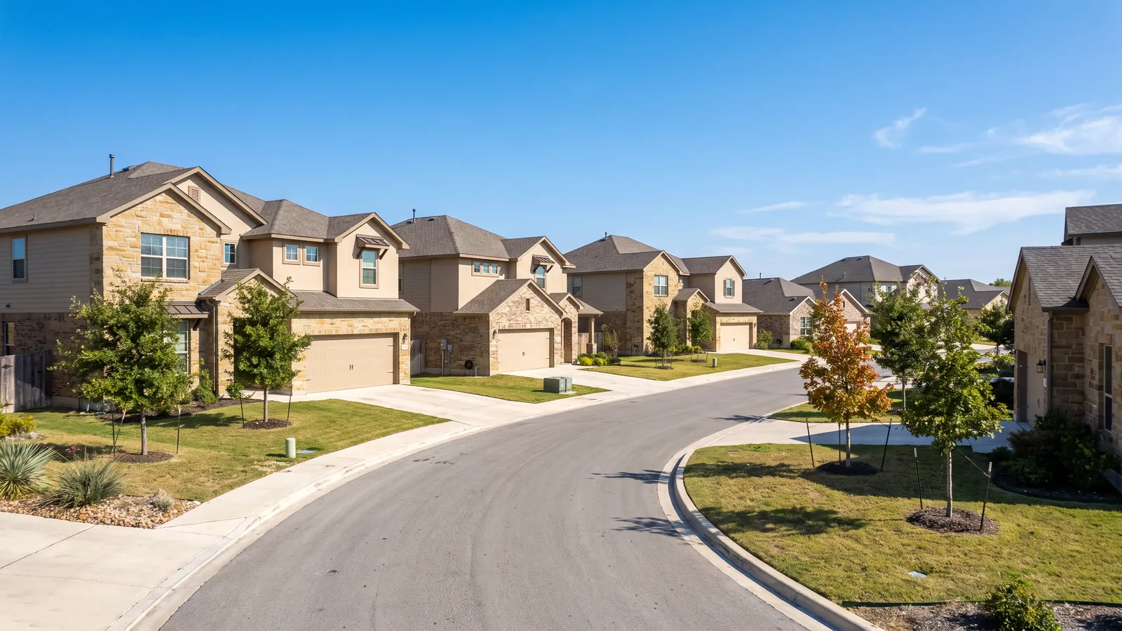 Curving subdivision street of newer two-story stone-and-stucco homes with front-facing two-car garage doors in the northern Austin suburbs, bright Texas sky and young trees