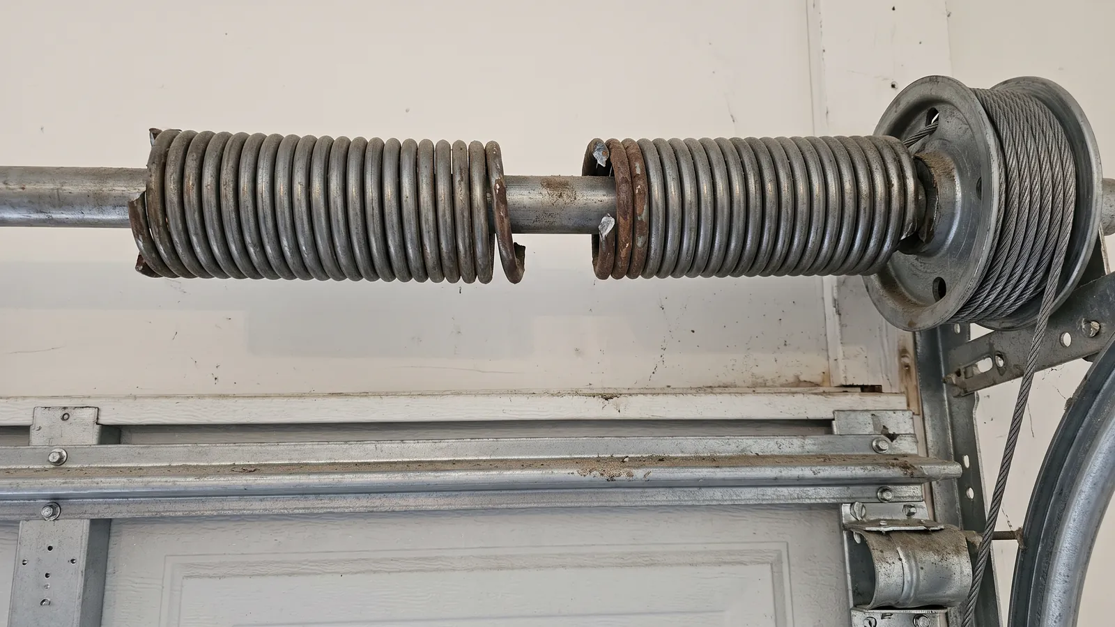 Close-up of a torsion spring that has snapped in two on the shaft above a steel sectional garage door, a clear gap between the broken coil ends and the cable drum to the right